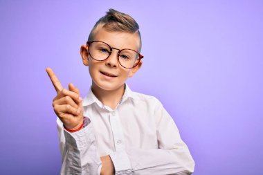 Mavi gözlü, mor arka planda beyaz gömlekli, yüzünde kocaman bir gülümseme olan, elleri ve parmaklarıyla kameraya bakan küçük beyaz bir çocuk..