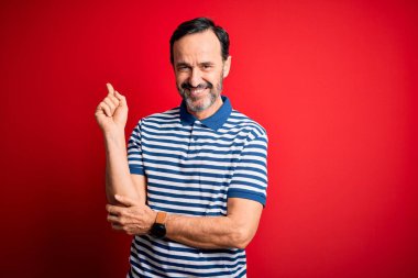 Orta yaşlı, beyaz bir adam, sıradan çizgili polo giyen, kırmızı arka planda duran, yüzünde kocaman bir gülümsemeyle, eli ve parmağı kameraya bakan,.