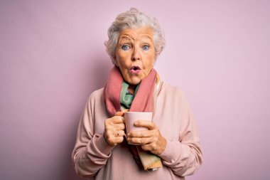 Son sınıf güzeli, gri saçlı bir kadın izole edilmiş pembe arka planda kahve fincanı içerken şaşkınlık içinde, korkmuş ve korku ifadesinden heyecanlanmış.