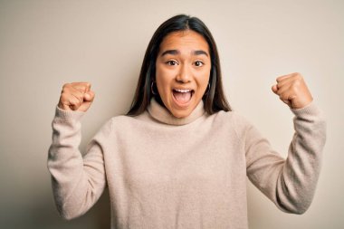 Beyaz arka plan üzerine balıkçı yaka kazak giyen genç, güzel Asyalı kadın kollarını kaldırıp, gözlerini açarak başarının şaşkınlığını ve şaşkınlığını yaşıyor. Kazanan konsept.