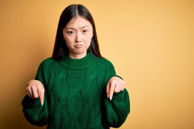 Sarıya boyanmış yeşil süveter giyen genç Asyalı kadın üzgün ve üzgün görünüyor, parmaklarıyla yönü gösteriyor, mutsuz ve depresif..