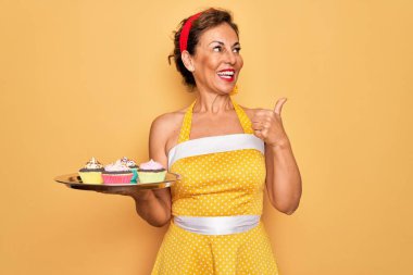 Orta yaşlı, son sınıf rozetli, 50 'li yılların modasına uygun elbise giyen, elinde kek tepsisi olan mutlu yüzlü bir kadın.