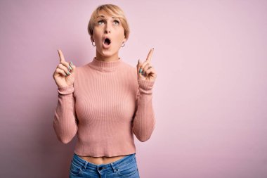 Kısa saçlı, balıkçı yaka kazak giyen genç sarışın kadın pembe arka planda şaşkın şaşkın yukarı bakıyor, parmaklarıyla işaret ediyor ve kollarını kaldırıyordu..