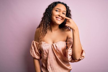 Kıvırcık saçlı, pembe arka planda sıradan tişörtler giyen, el parmağı ile yüzünü ve burnunu gösteren, neşeli gülümseyen güzel bir kadın. Güzellik konsepti