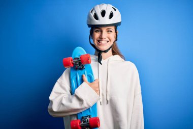 Güvenlik kaskı takan genç, güzel, kızıl kaykaycı kadın mutlu bir yüzle kaykay tutuyor ve gülümseyerek dişlerini gösteriyor.