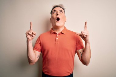 Soyutlanmış arka planda duran yakışıklı modern adam şaşırmış ve şaşırmış bir şekilde yukarı bakıyor ve parmaklarıyla işaret ediyor ve kolunu kaldırıyor..