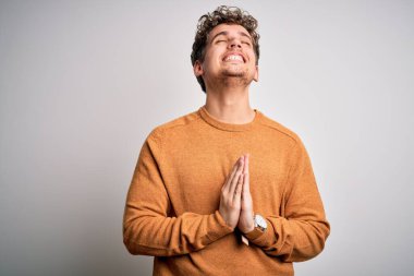 Sarışın, kıvırcık saçlı, beyaz arka plan üzerine gündelik kazak giyen, elleri bağlı, yalvaran ve dua eden, yüzünde umut ifadesi olan genç, duygusal ve endişeli bir adam. Yalvarıyorum..