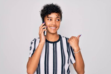 Genç, güzel, Afro-Amerikan bir kadın akıllı telefondan konuşuyor ve gülümseyen bir gülümsemeyle baş parmağıyla işaret ediyor.