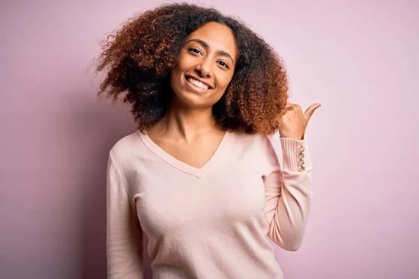 Young african american woman with afro hair wearing casual sweater over pink background smiling with happy face looking and pointing to the side with thumb up.