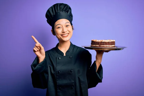 若い中国人シェフの女性を身に着けている炊飯器の制服と帽子を保持トレイケーキとともに驚きでアイデアや質問を指差す指で幸せな顔 ナンバーワン — ストック写真