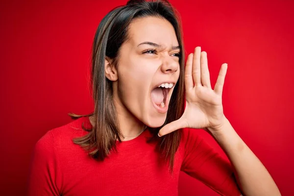若い美しいブルネットの女の子は 孤立した赤い背景の叫びの上にカジュアルなTシャツを着て 口の上に手で横に大声で叫んでいます コミュニケーションの概念 — ストック写真