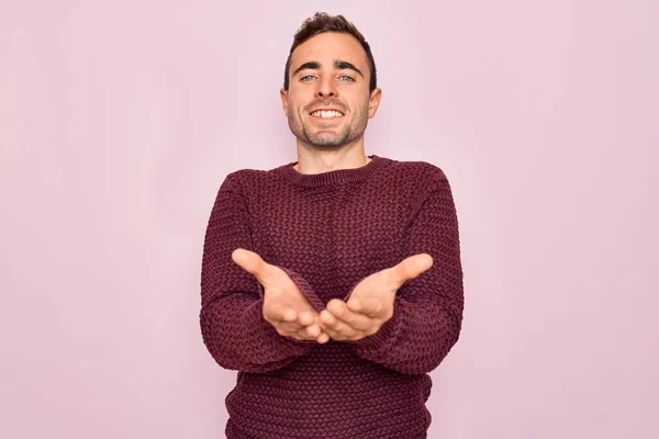 青い目をした若いハンサムな男がピンクの背景に立ってカジュアルなセーターを着て一緒に手の手のひらで笑顔を受信またはジェスチャーを与える ホールドと保護 — ストック写真