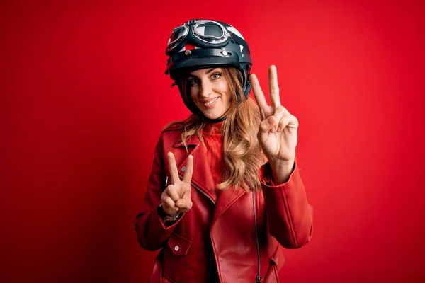 若い美しいブルネットのオートバイの女性は 勝利のサインをして指を示すカメラを見て笑って赤い背景の上に元ヘルメットを着ていました 第二番 — ストック写真