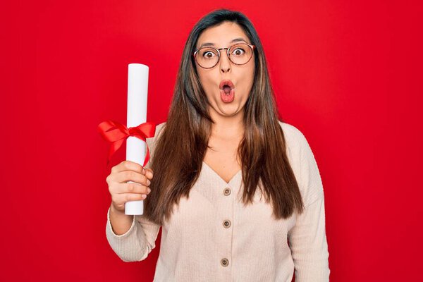 Young hispanic smart woman wearing glasses holding university degree over red background scared in shock with a surprise face, afraid and excited with fear expression