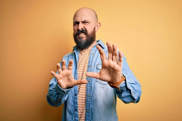 Handsome bald man with beard wearing casual denim jacket and striped t-shirt disgusted expression, displeased and fearful doing disgust face because aversion reaction. With hands raised