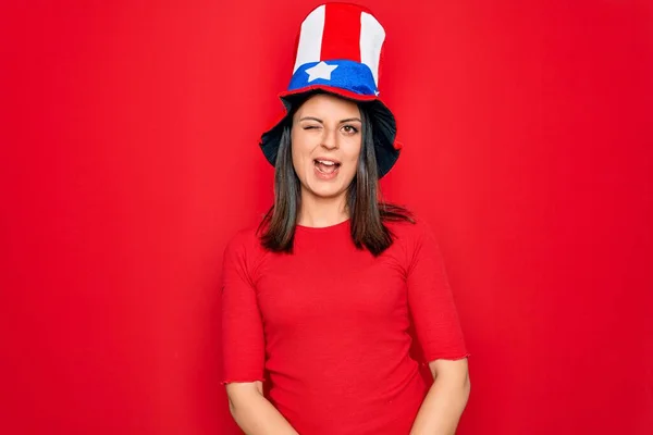 Young beautiful brunette woman wearing united states hat celebrating independence day winking looking at the camera with sexy expression, cheerful and happy face.