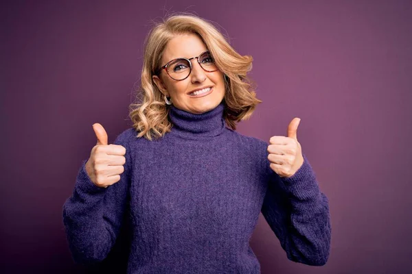 Middle age beautiful blonde woman wearing casual purple turtleneck sweater and glasses success sign doing positive gesture with hand, thumbs up smiling and happy. Cheerful expression and winner gesture.