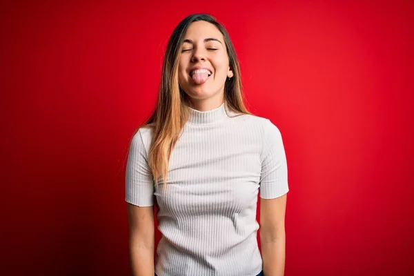 Beautiful blonde woman with blue eyes wearing casual white t-shirt over red background sticking tongue out happy with funny expression. Emotion concept.