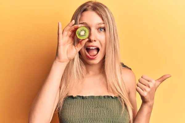 Young Blonde Woman Holding Kiwi Pointing Thumb Side Smiling Happy — ストック写真