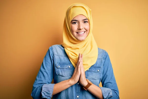 Young beautiful girl wearing muslim hijab standing over isolated yellow background praying with hands together asking for forgiveness smiling confident.