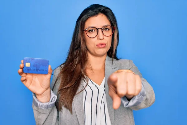 Young hispanic business woman holding credit card over blue isolated background pointing with finger to the camera and to you, hand sign, positive and confident gesture from the front