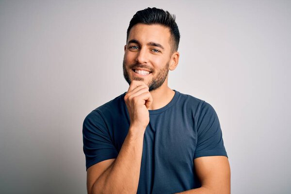 Young handsome man wearing casual t-shirt standing over isolated white background looking confident at the camera with smile with crossed arms and hand raised on chin. Thinking positive.