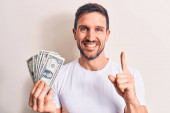 Mladý pohledný muž držící hromadu dolarů bankovek nad izolovaným bílým pozadím, usmívající se nápadem nebo otázkou ukazující prstem se šťastnou tváří, číslo jedna