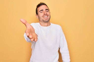 Mavi gözlü, sıradan bir süveter giyen, sarı arka planda gülümseyen, yardım eli uzatan ve kabul gören genç yakışıklı bir adam..