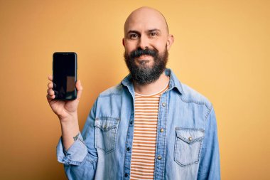 Sakallı yakışıklı kel bir adam. Akıllı telefon tutuyor. Sarı arka planda gülümseyen bir yüz ve kendine güvenen bir gülümsemeyle gülümsüyor.
