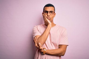 Yakışıklı Afro-Amerikalı adam pembe arka plan üzerinde günlük tişört ve gözlük takıyor. Kollarını kavuşturup depresyondan sıkılmış ve yorgun görünüyor..