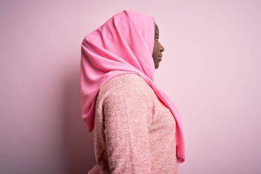 Genç Afro-Amerikan artı cüsseli kadın soyutlanmış pembe arka plan üzerine Müslüman başörtüsü takıyor. Yan tarafa bakıyor, rahat poz veriyor, kendine güvenen bir gülümsemeyle doğal bir görünüm sergiliyor..