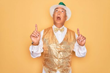 Sarı arka planda Brezilya karnavalı kıyafeti giyen orta yaşlı, kır saçlı bir adam şaşırmış ve şaşırmış bir şekilde yukarı bakıyor ve parmaklarıyla işaret ediyor ve kollarını kaldırıyor..