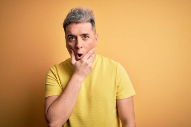 Sarı izole edilmiş arka plan üzerinde sarı tişört giyen genç, yakışıklı modern adam elleri çenesinde olan inançsızlığa, şaşkınlığa ve hayrete düşmüş bir ifadeye bakıyor.