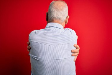 Orta yaşlı, beyaz, sıradan çizgili gömlek ve kırmızı arka planda gözlük takan yakışıklı bir adam mutlu ve pozitif bir şekilde kendine sarılıyor. Sevgi ve özgüven.