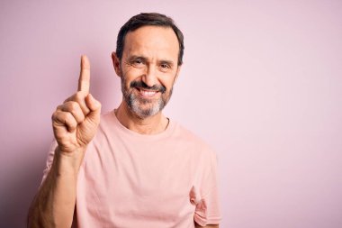 Orta yaşlı, beyaz tişörtlü, izole edilmiş pembe arka planda duran ve bir numarayı işaret eden kendinden emin ve mutlu bir şekilde gülümseyen bir adam..