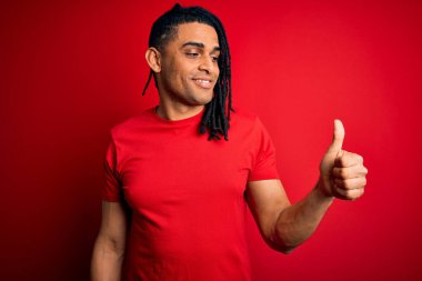 Kırmızı günlük tişört giyen, gururlu, gülümseyen, baş parmak hareketi yapan, rastalı saçlı Afro-Amerikan genç adam.