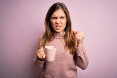 Genç sarışın kadın pembe izole edilmiş arka planda bir fincan kahve içiyor sinirlenmiş ve hayal kırıklığına uğramış öfkeyle bağırıyor, deliye dönmüş ve el kaldırarak bağırıyor.