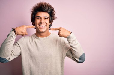 İzole edilmiş pembe arka plan üzerinde sıradan tişörtler giyen genç ve yakışıklı bir adam gülümseyip, parmaklarını ve ağzını işaret ediyor. Diş sağlığı konsepti.