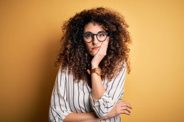 Kıvırcık saçlı, piercingli, çizgili tişörtlü ve gözlüklü genç bir kadın kollarını kavuşturup depresyondan sıkılmış ve yorgun göründüğünü düşünüyor..