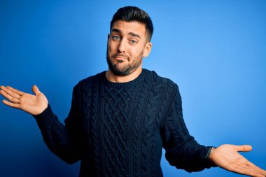 İzole edilmiş mavi arka plan üzerinde duran sıradan bir süveter giyen genç ve elleri ve kolları havada, kafası karışmış bir adam. Şüphe kavramı.