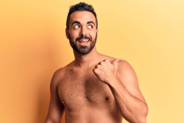 Genç İspanyol adam gülen yüzüyle üstsüz duruyor başparmağıyla yan tarafa bakıyor ve işaret ediyor.. 