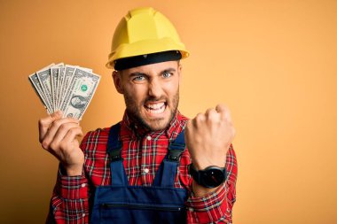 Güvenlik kaskı takan genç müteahhit, elinde dolar tutarak sarı arka plan ödemesi yapıyor. Sinirli ve sinirli bir şekilde bağırıyor, çıldırıyor ve el kaldırarak bağırıyor. Öfke kavramı.
