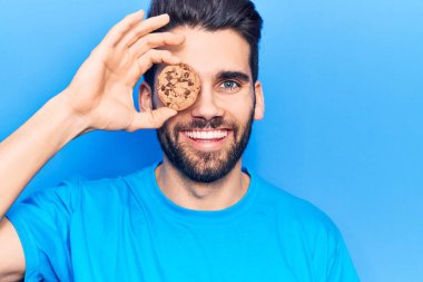 Sakallı, elinde çikolatalı kurabiyeyle genç ve yakışıklı bir adam pozitif ve mutlu görünüyor ve kendine güvenen bir gülümsemeyle dişlerini gösteriyor. 