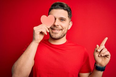 Mavi gözlü genç romantik bir adam kırmızı arka planda kırmızı renkli kağıtlar tutuyor. Bir fikir ya da soru işareti ile şaşırıyor. Mutlu yüzle işaret ediyor.