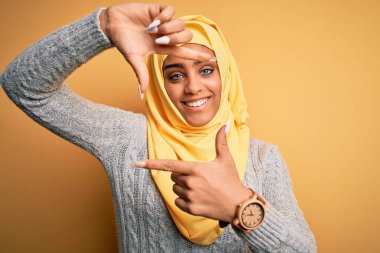 Genç ve güzel Afro-Amerikalı kız izole edilmiş sarı arka plan üzerine Müslüman başörtüsü takıyor gülümsüyor ve elleri ve parmakları mutlu yüzle çerçeve yapıyor. Yaratıcılık ve fotoğrafçılık kavramı.