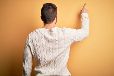 Sarışın, sakallı, mavi gözlü, sarı arka planda beyaz süveter giyen genç bir adam.