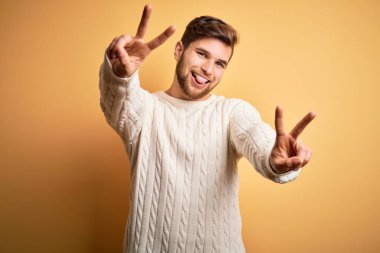 Sarışın, sakallı, mavi gözlü, sarı arka planda beyaz süveter giyen, iki elini de zafer işareti yaparken gösteren bir dili olan genç bir adam. İki numara..