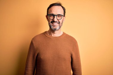 Orta yaşlı, beyaz, kahverengi süveter ve gözlük takan mutlu ve havalı bir gülümsemesi olan izole edilmiş sarı arka plan. Şanslı kişi..