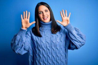 Mavi gözlü genç esmer kadın balıkçı yaka kazak giyiyor ve parmaklarıyla on numarayı gösterirken kendinden emin ve mutlu bir şekilde gülümsüyor..