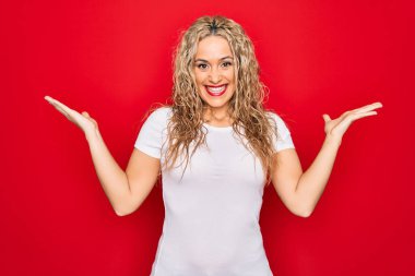 Genç ve güzel sarışın bir kadın, gündelik tişört giyip izole edilmiş kırmızı arka planda dikilmiş çılgınlığı kutluyor ve başarıya hayret ediyor. Kolları havada ve gözleri açık, heyecanlı bir şekilde çığlık atıyor. Kazanan konsept